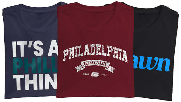 Banner featuring three folded t-shirts with Philadelphia themes: one with 'IT'S A PHILLY THING' in teal and navy, a maroon one with 'PHILADELPHIA Pennsylvania ESTD 1682,' and a black one with 'jawn' in red and blue.