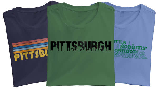 Banner showing three folded t-shirts with Pittsburgh themes: navy with a sunset and skyline, green with a dripping skyline, and blue with 'Mister Rogers' Neighborhood' text.