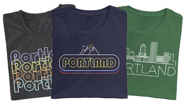 Banner displaying three folded t-shirts with Portland designs: charcoal with multicolored text overlay, navy with a retro 'PORTLAND' logo, and green with a white cityscape outline.