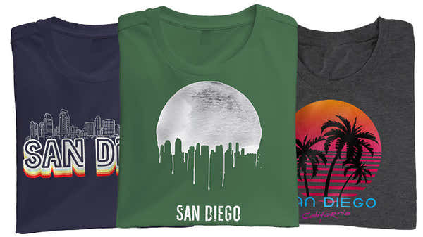 Banner featuring three folded t-shirts with San Diego designs: navy with colorful skyline and '61', green with a white dripping moon, and charcoal with a sunset and palm trees.