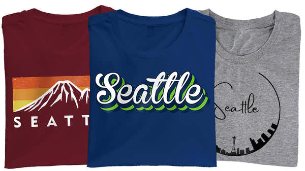 Banner displaying three Seattle-themed t-shirts: maroon with a mountain sunset and 'Seattle' text, blue with stylized 'Seattle' in green and white, and grey with 'Seattle' encircled by city landmarks.