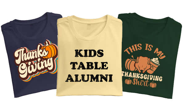 Three Thanksgiving-themed t-shirts; navy with 'Thanksgiving' and a pumpkin, cream with 'KIDS TABLE ALUMNI' in black lettering, and green with 'THIS IS MY THANKSGIVING SHIRT' with a turkey graphic.